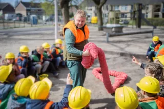 Kinderen zitten met een bouwhelm op in een kring op het schoolplein. In het midden staat een acteur met een grote wollen roze worm.